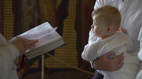 Child boy in the Orthodox Church at the baptism ceremony. Christening baby, Catholic or Christian religion. Religious rite. Godfather and Godmother. Priest reads prayer from bible book or gospel book.