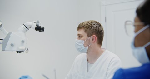 A male dentist of European appearance looks through a dental microscope for the treatment of a patient's tooth root canal. Removal of a nerve in a tooth with a modern method.