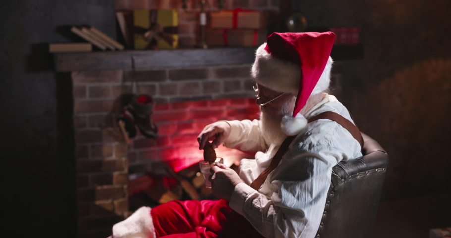 Happy Santa Claus eating ginger cookies, sitting near burning fireplace. Mr Claus free time, relaxing after work in cozy house residence. Post office of Santa, leisure. Christmas atmosphere, New Year.
