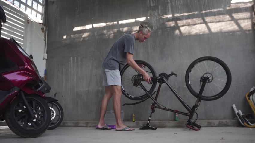 Young man with hair bun spins bicycle pedal with hand to check sports bike wheel standing in garage against grey wall and scooters Royalty-Free Stock Footage #1080666146