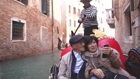 Wide shot of a beautiful mature married couple rides on a gondola boat in typical Venetian canals and takes a video call or selfies in Venice, in autumn, or winter. Italian gondolier. Go everywhere.