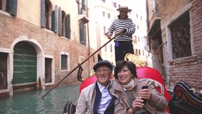 Wide shot of a beautiful mature married couple rides on a gondola boat in typical Venetian canals and takes a video call or selfies in Venice, in autumn, or winter. Italian gondolier. Go everywhere.