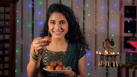 A young woman offering homemade Gulaab Jamun to her guests on Diwali. Beautiful brass Diyas burning brightly against the festive background - a Hindu festival, an auspicious occasion, positive vibes