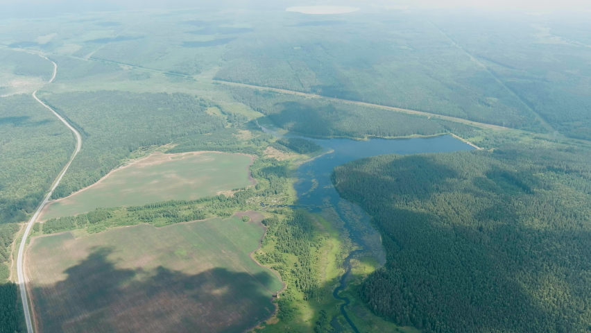 Russia, Ural. Aerial ascent over a vast expanse of mixed boreal forest, open meadows, and a winding swampy pond, with long shadows of clouds drifting across the terrain. The camera rises steadily through a layer of soft atmospheric haze, revealing the flat-to-rolling topography characteristic of the West Siberian Plain at the eastern edge of the Ural range. A straight road cuts through the landscape on the left, providing scale against the dense green canopy and the reflective water body below. The scene captures the transition from clear summer daylight into a milky, cloud-diffused sky, emphasizing the remote and untouched nature of this wetland ecosystem. The Ural Mountains, among the world