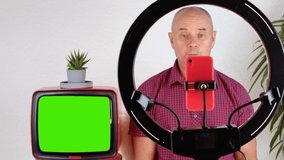 middle-aged charming man, 60-65 years old blogger sits in front of a ring light and a red smartphone, emotionally talks on the topic of business, politics, old TV with a green screen for video