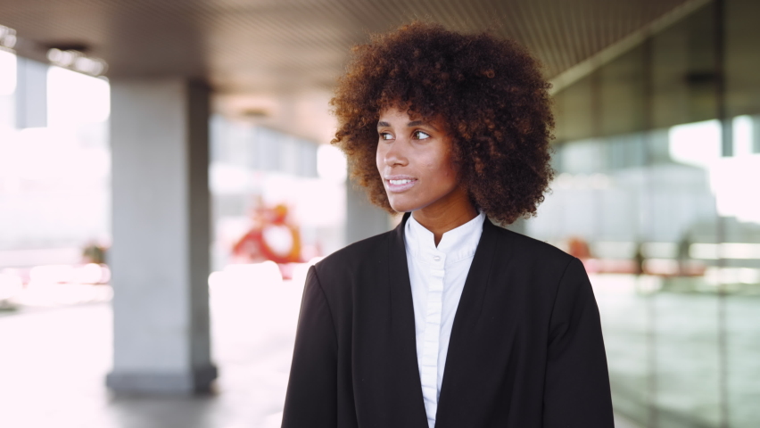 Medium Slow Motion Tracking Shot Of Young Businesswoman With Afro Hair Breaking Into Toothy Smile To Camera As She Walks Along