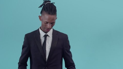 Tired black office worker in a suit takes hand tie and simulates hanging with tongue stuck out in the studio blue background. Black male businessman with dreadlocks shows a noose out of tie. Fatigue