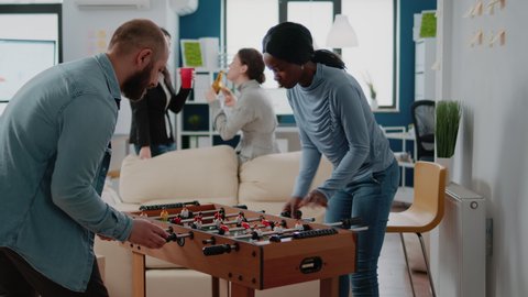 Man and woman playing football game at foosball table to celebrate party after work. Group of workmates enjoying play and drinks while eating pizza after hours at office. Cheerful coworkers