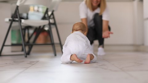 Cute newborn baby girl in white body growling on white warm floor to her mom at home. Focus is at feet. 