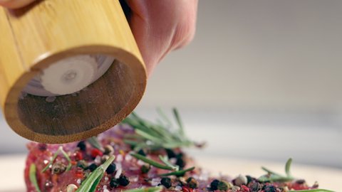 Person grinding peppercorns onto raw meat for seasoning in a close up view using a wooden pepper mill with fresh herbs and spices in a cookery concept