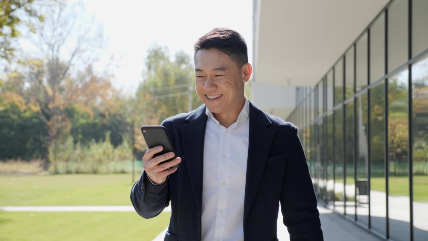 Portrait of asian successful businessman wearing stylish suit and shirt walking outdoor near the office buildings. Man smiling while holding smartphone outdoor. Business lifestyle concept. Royalty-Free Stock Footage #1080690578