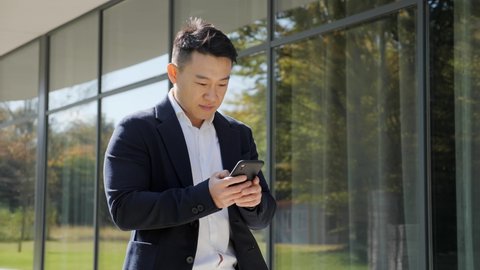 Attractive asian businessman enjoying successful startup while looking on the screen of his smartphone. Handsome korean shocked male received positive news and showing yes gesture. Success concept.