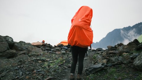 A woman in the mountains climbs up with a large backpack with equipment and poles for a hike came to the camp with tents. Climber in the rocks walks the trail while hiking