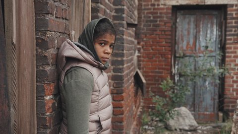Medium portrait of abandoned 11-year-old African-American girl looking at camera standing outdoors against brick wall at poor district