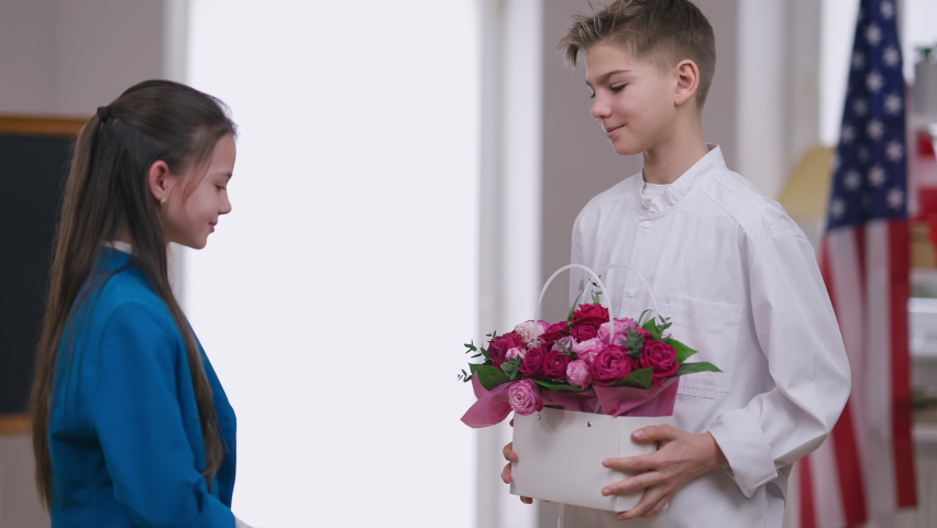 Smiling boy in doctor gown congratulating girl on Women's Day surprising sister with basket of flowers. Thankful Caucasian sibling hugging brother talking. Family unity and holiday celebration