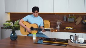 Smiling asian young man study play guitar by video call with teacher. Home kitchen, distance learning lesson with smartphone. Kind friendly pupil person. Slow motion, Copy space, for text