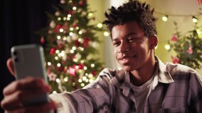 African American man speaking on video call on smartphone, sitting on sofa. Black afro guy talking with friends online in app. Christmas time concept. New Year holidays. Cozy winter evening at home.