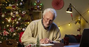 Old man pensioner teacher writing notes close-up. Grandfather in glasses, speaking on video call on laptop, sitting in decorated living room. Christmas holidays concept. New year and winter time. 