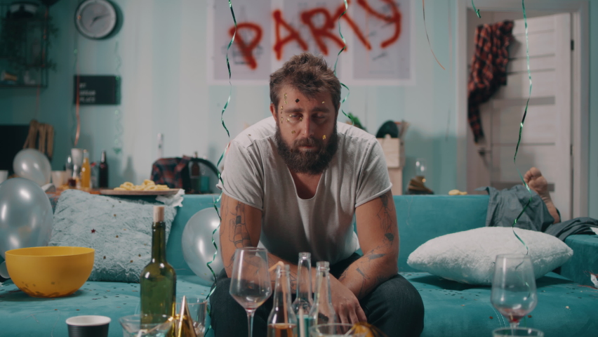 Zoom out view of bearded man with hangover touching face and spitting out confetti while sitting on sofa in messy living room after party Royalty-Free Stock Footage #1080712214