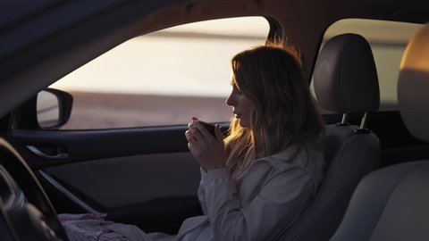 Woman inside the car is drinking coffee from the thermos cup, adoring sunrise on the seaside