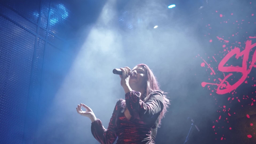 A female performer wearing a red dress singing into a cordless microphone against a blue stage with a spotlight shining from above in a smoke-filled room. Royalty-Free Stock Footage #1080724910