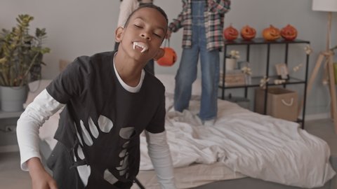 Slowmo tracking shot of 12-year-old African-American boy in Dracula costume and vampire teeth making spooky faces om camera while his friends jumping on bed during Halloween home party