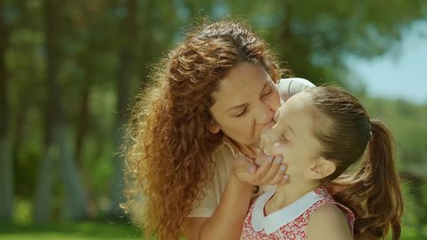 Beautiful mother and her beautiful little daughter are having fun together on the grass outdoors. Mom kisses her little daughter on the cheek. Happy Mother's Day. 