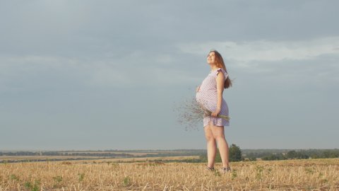 camera rotating around happy pregnant woman in dress standing alone in field with a bouquet of wild flowers on background of cloudy rainy sky, relaxation and healthy pregnancy concept