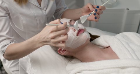 Face mask, beauty treatment, skincare. Young caucasian female getting facial nourishing mask by confident professional beautician at spa salon, side view, close-up. lady is relaxed lying on couch