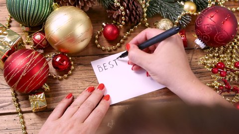Woman's hand writing BEST card on the wooden table with Christmas decorations around closeup	