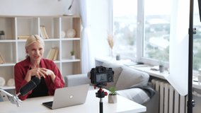 Video blog. Business coaching. Virtual training. Cheerful inspired senior influencer woman speaking recording content on camera on tripod on defocused light home interior background.