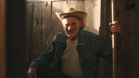 Positive elderly man with tool smiling and looking at camera while sitting near lumber wall of stable during work on farm