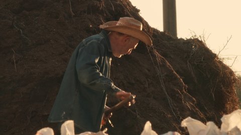 Elderly male farmer in casual clothes and straw hat filling bags with soil while working on ranch in summer