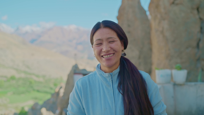 A bright and smiling middle aged Asian adult female with long black hair standing outdoors with snow-clad mountains in the back looking at the camera Royalty-Free Stock Footage #1080766745