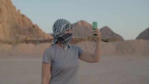 Female tourist in checkered keffiyeh trying to find mobile network using smart phone standing in desert. Woman searching for mobile signal to make a call in desert. 