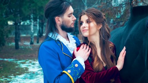 Beautiful fabulous man in love tenderly kisses happy redhaired woman. Romantic meeting of lovers in winter forest. Black horse. Blue vintage carnival camisole. Darkhaired medieval king, beard on face 
