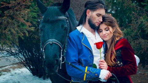 Closeup portrait loving family medieval couple. Prince man gently hugs fantasy fairytale woman princess. Redhaired lady holding red rose, enjoy winter nature forest park, smiling happily. Black horse 