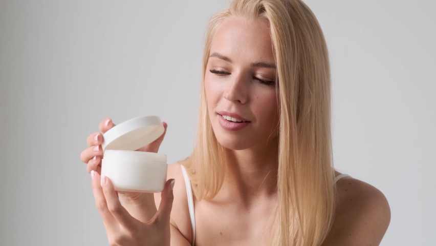 A smiling young blonde woman sniffs the scent of cream standing isolated over grey wall in the studio