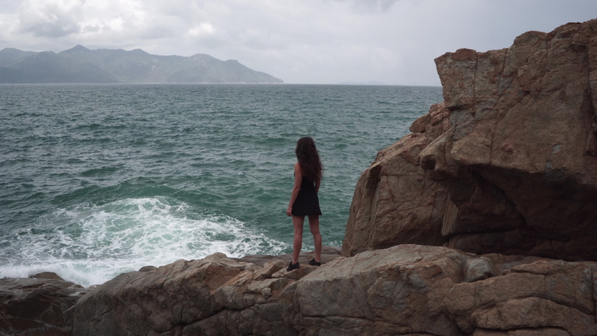 Woman traveler with dark hair in dress looks at azure sea waves crashing on beach cliff rocks on cloudy day backside view Royalty-Free Stock Footage #1080810149