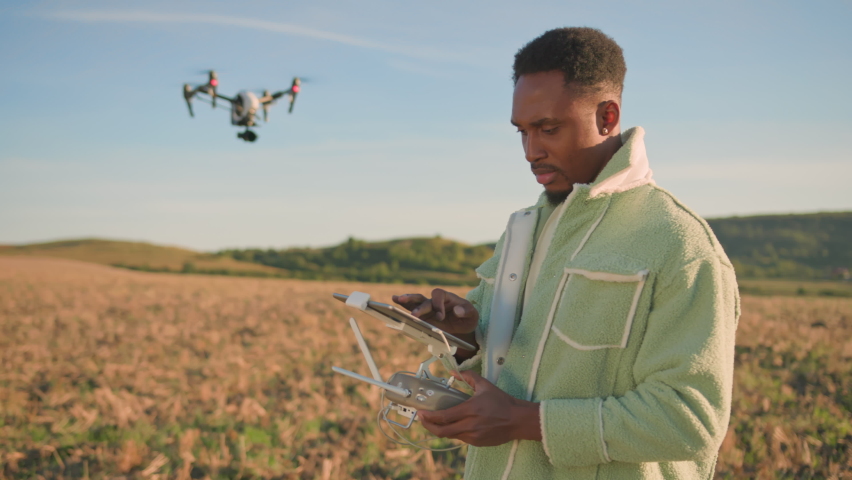 African american young farmer holding computer notepad for controlling drone flight. Futuristic black copter flying over field to monitor situation. Farming technology. Royalty-Free Stock Footage #1080814085