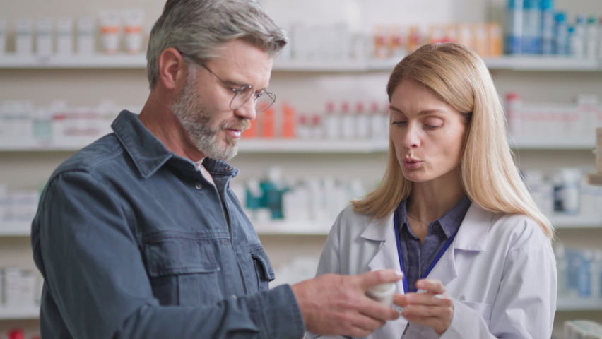 Caucasian adult man wearing glasses visiting drugstore, talking to female consultant about antiviral medicine or vitamins. Modern apothecary. Pharmacy. Royalty-Free Stock Footage #1080814199