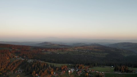 Aerial flight over hilly landscape lit with warm morning sunlight. Drone shot small villages, hills covered with pine trees in dawn haze orange sunrise light. Picturesque Zakopane village from above