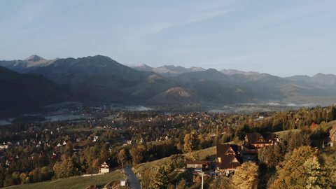 Aerial view flight over mountain village lit with warm sunlight. Drone shot beautiful highland valley in sunset light. Picturesque Tatra mountains range covered with clouds, Poland, Zakopane