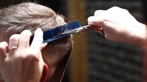 Professional unisex hairdresser with neat manicure cuts man short dark hair on back using steel scissors and comb in salon closeup