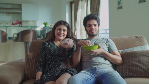 A young modern attractive couple is jumping or sitting on the couch and turn on the TV to watch a movie or cricket match. A girlfriend and boyfriend spending quality time together in an interior house