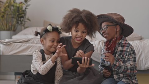 Slowmo shot of two African-American girls and boy in Halloween costumers making spooky faces while taking selfie portrait on camera indoors