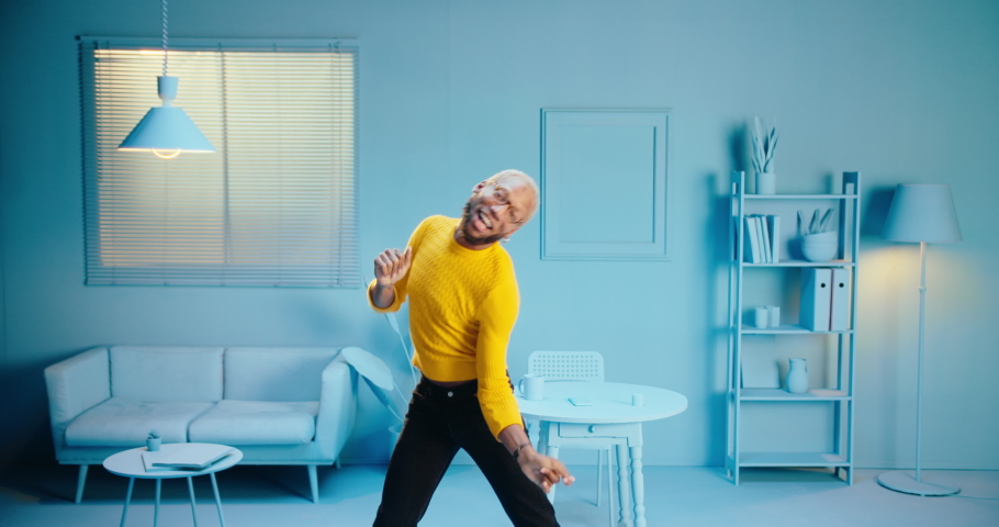 Excited cheerful black man having fun alone listening music and dancing at home with blue color decoration background, carefree African American person dancing with silly movements at modern home Royalty-Free Stock Footage #1080826322