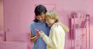 Portrait of two excited mixed race young girls using mobile phone laughing and hugging together at pink studio. Carefree two women reading message on a smart home at home with monochrome interior