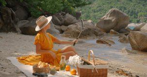 Pretty woman in yellow dress and straw hat having video call on summer day sitting on lonely beach
