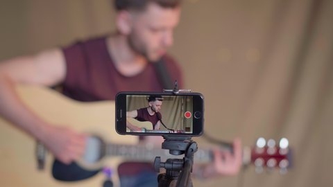 Man recording guitar lessons for students. Online guitar courses. Distance learning concept Caucasian male hipster playing acoustic guitar on video call Medium shoot Blogger teaching people guitar 